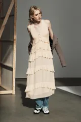 Vestido midi de escote en pico con lazada frontal y diseño sin mangas. Presenta múltiples niveles de volantes a lo largo de la falda y cierre lateral con cremallera oculta.