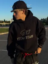 Hoodie de color negro con diseño de estampa frontal que combina una tipografía manuscrita con el texto ITTAI STARS en letras mayúsculas. Presenta un corte holgado, capucha amplia y bolsillo tipo canguro.