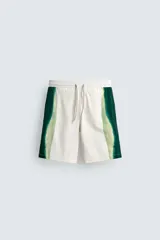 Bañador de hombre color blanco con diseño de bandas verticales en tonos de verde, simulando un efecto tie-dye o acuarela. Posee cintura ajustable con cordón, bolsillos laterales y forro interior.