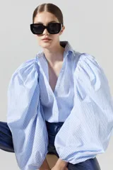 Camisa de algodón a rayas verticales blancas y celestes, con mangas globo muy voluminosas y puños elásticos. Posee cuello camisero clásico y cartera de botones de nácar. El ruedo es más largo en la parte trasera.