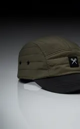 Gorra de estilo 5 paneles, color verde militar, con corona acolchada y visera negra. Presenta un pequeño parche negro con un logo blanco en forma de tijeras cruzadas.