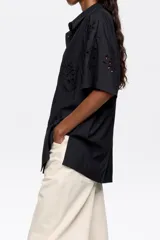 Camisa negra de manga corta con cuello clásico y cierre frontal con botones. Presenta bordado inglés con motivos florales. Tiene un bolsillo de parche en el pecho.