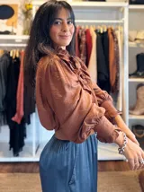 Blusa de color marrón con diseño romántico, cuello alto con lazo ajustable y volados tipo shabot en el frente. Presenta mangas largas abullonadas con puños ajustados y una textura suave y liviana.