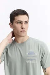 Remera de manga corta color verde salvia, con escote redondo y fit regular. Presenta un estampado frontal con texto en tonos azul y blanco, incluyendo la marca Mistral y el número "19SEVENTYSIX".