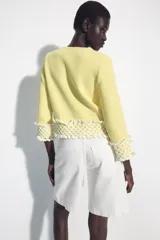 Chaqueta de punto color amarillo claro, con cuello redondo y mangas por debajo del codo. Presenta detalles de acabado en tejido crochet blanco en el ruedo y los puños, con cierre frontal de botones forrados al tono.