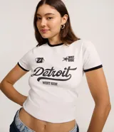 Remera ajustada de manga corta color blanco con ribetes negros en cuello y mangas. Presenta un estampado frontal con la palabra "Detroit" en tipografía cursiva grande, y texto más pequeño que dice "WEST SIDE" debajo. También incluye el número "23" y un logo con una estrella y el texto "WST SIDE".