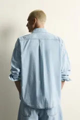 Camisa de corte relajado confeccionada en denim ligero de algodón, con cuello solapa, manga larga con puños abotonados y bolsillos de plastrón en el pecho. Presenta un acabado con efecto lavado y cierre frontal mediante botones.