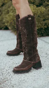 Bota alta de gamuza marrón con caña desmontable para transformarla en bota corta. Presenta detalles de tachas decorativas, puntera cuadrada y taco ancho.
