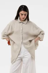 Camisa de lino color beige, de calce amplio y relajado, con cuello camisero, cierre frontal de botones, mangas largas y diseño de largo irregular con aberturas laterales.