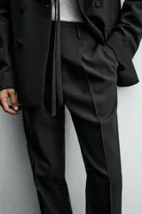Pantalón de vestir negro con pliegues frontales, corte regular fit, bolsillos laterales y traseros de doble vivo con botón. Cierre frontal cruzado con cremallera y gancho metálico.