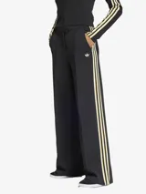 Pantalón de corte holgado y estilo sastrero, confeccionado en tejido de doble punto color negro. Presenta las icónicas tres tiras de la marca en los laterales en combinación de colores blanco y amarillo, con el logo del trébol bordado en la parte frontal. Cuenta con cierre de cremallera y bolsillos laterales.