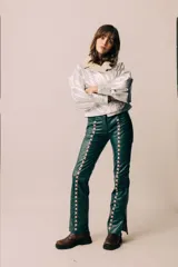 Pantalón de cuero verde oscuro, de tiro medio y silueta recta levemente acampanada, con recortes laterales intervenidos con pasacintas entrelazadas en contraste y tajo en los tobillos.