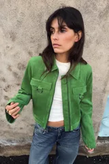 Chaqueta corta de tejido efecto ante en color verde vibrante, con cuello solapa, cierre frontal de cremallera y mangas largas. Presenta dos bolsillos frontales con solapa decorados con arandelas metálicas en el pecho.