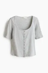 Top de manga corta de punto pointelle suave, color gris, con ajuste entallado, escote en U y hilera de botones frontales. Presenta ribete levemente festoneado en el escote y la tapeta.