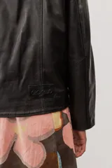 Chaqueta de cuero negra con cuello redondo, cierre frontal de botones forrados y bolsillos ribeteados con detalles de costura decorativa.