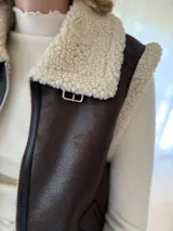 Chaleco de gamulán color marrón con interior y bordes de corderito sintético color crema. Presenta cierre frontal metálico y diseño sin mangas, ideal para superponer sobre otras prendas.