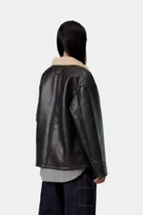 Chaqueta de trabajo Carhartt WIP modelo Michigan Shepton, color negro, confeccionada en piel sintética con cuello y forro de corderito sintético en tono tostado. Presenta cierre frontal de botones, cuatro bolsillos delanteros de parche (uno con solapa) y etiqueta cuadrada con el logo de la marca.