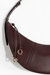Cartera marrón de diseño curvo con costuras visibles, correa ajustable, cierre superior y tiradores largos con detalle de nudo y argollas doradas.