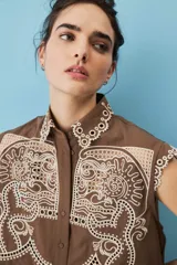 Camisa cropped sin mangas color tostado, con cuello clásico, cierre frontal de botones, bordado calado y terminación festoneada en el ruedo.