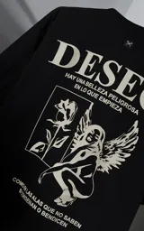 Remera de manga corta y cuello redondo con calce oversize. Presenta una estampa frontal en color crema con diseño artístico de un ángel y una rosa, acompañada de frases en español. Confeccionada en algodón premium.
