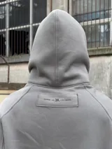 Campera con capucha gris oscura, cierre frontal y diseño oversize con doble capucha, incluyendo balaclava integrada. Presenta gran estampa contrastante en el frente.