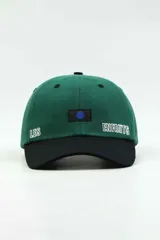 Gorra con visera de diseño bicolor, con cuerpo en color verde y visera en color negro. Presenta un parche rectangular con un punto azul en el centro y la palabra 'ENFANTS' bordada en blanco en el panel frontal.