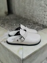 Zuecos blancos de cuero sintético con hebilla metálica ajustable.