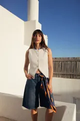 Chaleco sastrero blanco sin mangas, con cuello redondo y cierre frontal de botones con efecto carey. Presenta bolsillos ojal simulados en la parte inferior.