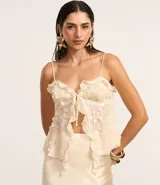 Blusa corta color crema con escote corazón, confeccionada en tejido con textura de lunares y volados de chifón. Presenta breteles finos ajustables y un lazo decorativo en el centro del busto.