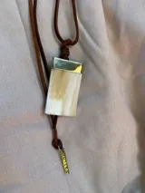Collar largo con tiras de cuero marrón y dije rectangular de nácar con aplique superior metálico dorado y placa colgante grabada.