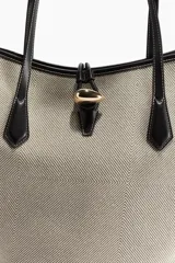Cartera tipo shopper con diseño de borde superior ondulado y estructura sesgada. Presenta asas finas de cuero negro y un cierre central con detalle metálico dorado. El cuerpo de la cartera posee un patrón geométrico sutil en tonos beige y gris.