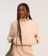 Campera bomber color beige confeccionada en terciopelo cotelé, con cuello alto abotonado, cierre delantero con botones, bolsillos laterales, mangas largas ajustadas y bajo ajustado con elástico.