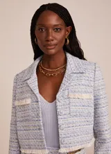 Chaqueta corta de tweed celeste con detalles en blanco y dorado, de corte sastre, con solapas de pico, cierre frontal con botones, bolsillos delanteros y terminaciones con flecos.