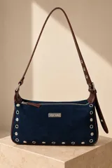 Cartera tipo bandolera de gamuza azul marino con ribetes y correa de cuero marrón. Presenta tachas y ojalillos metálicos a lo largo de los bordes frontales, cierre superior y una placa metálica con el logo de la marca en el centro.