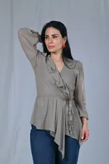 Blusa color gris beige con escote cruzado en V y volados en el frente. Tiene mangas largas con puños elásticos y corte a la cintura con lazo lateral.
