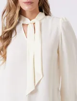 Blusa de gasa color crema, marca Zac & Rachel, con escote en V y lazo para anudar en el cuello. Mangas largas con puños abotonados.