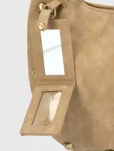 Cartera tipo hobo de Hush Puppies en color taupe, confeccionada en material con textura gamuzada. Presenta un diseño de media luna con tachas metálicas decorativas en los laterales y la base, asa única integrada y un dije colgante con el logo de la marca.