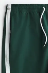 Short deportivo de color verde oscuro, confeccionado en nylon, con bandas laterales blancas, cintura elástica ajustable mediante cordón blanco y corte estándar.