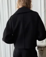 Chaqueta negra de corte corto y holgado, confeccionada con mezcla de lana. Cuenta con cuello ancho y solapa de piel sintética en tono beige claro, cierre frontal con botones oscuros y mangas largas amplias.