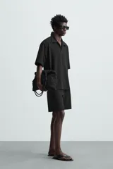 Bermuda de hombre color verde claro, de corte regular y tiro medio, con pinzas frontales, bolsillos laterales y bolsillos traseros de vivo. Cierre frontal con botón y cremallera.