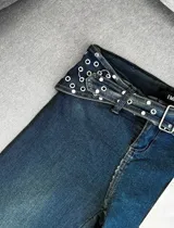 Pantalón jean de corte recto y tiro alto, color azul oscuro, con pretina ancha decorada con múltiples ojales metálicos y tachas, e incluye un cinturón incorporado con hebilla metálica.