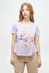 Remera de algodón color lila con escote en V y mangas cortas. Presenta un estampado central artístico que combina flores y elementos marinos. El diseño cuenta con un corte ligeramente más largo en la parte posterior.