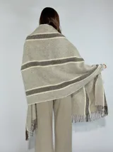 Ruana tipo pashmina color tostado con diseño a cuadros y rayas en tonos beige y marrón. Presenta flecos en los bordes.