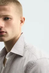 Camisa de corte slim fit confeccionada en tejido de algodón, con estampado de cuadros pequeños en tonos beige y blanco. Presenta cuello italiano, manga larga con puños abotonados y cierre frontal con botones.