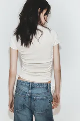 Camiseta corta de cuello redondo y manga corta, color gris oscuro, con frunces laterales ajustables mediante cordones.