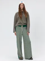 Pantalón de punto jacquard con diseño geométrico en tonos verde y beige. Presenta un corte holgado de pierna ancha, tiro medio, cintura elástica con cordón ajustable y franjas laterales en color verde sólido.