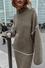 Jersey de punto con cuello subido y manga larga, de corte holgado. Presenta detalles de vivos en color crema en el cuello, puños y costuras de las mangas, contrastando con el tono marrón jaspeado de la prenda.