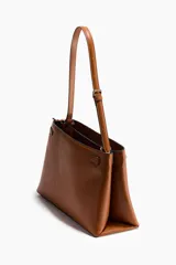 Cartera de hombro color tostado, fabricada en tejido revestido, con correa ajustable y un divisor interior que crea dos compartimentos, además de un compartimento interior con cierre. Presenta una trabilla decorativa con mosquetón metálico dorado en la parte delantera y una etiqueta extraíble.