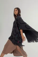 Ruana de tejido de punto grueso en color gris oscuro, con diseño de líneas curvas en relieve de color negro y detalles de costuras decorativas en colores vibrantes. Presenta terminaciones con flecos en el ruedo y una etiqueta de cuero sintético en la parte inferior.