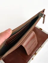 Billetera tipo porta celular de cuero, con diseño combinado en cuero liso marrón y cuero con pelo vacuno estampado. Cuenta con cierre superior, correa de muñeca y solapa frontal con cierre magnético y logo de búho grabado.